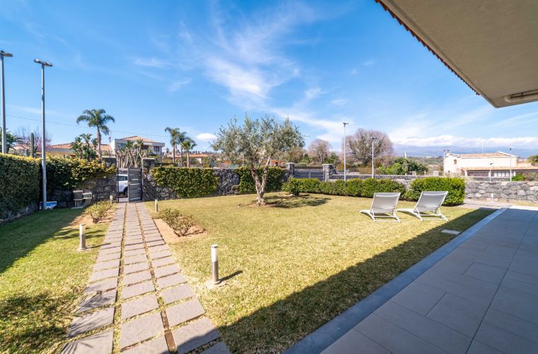 The exterior features a well-kept lawn and olive trees.